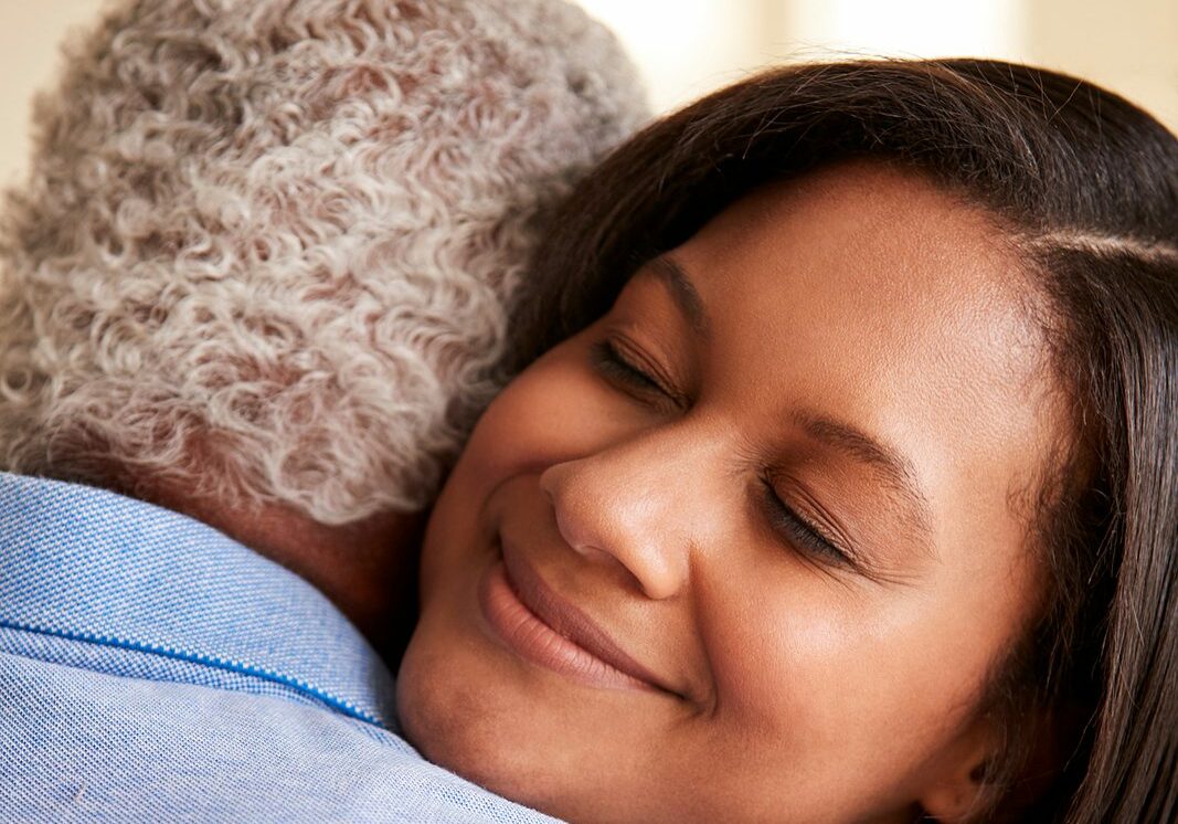 Over The Shoulder View Of Senior Father Being Hugged By Adult Daughter At Home