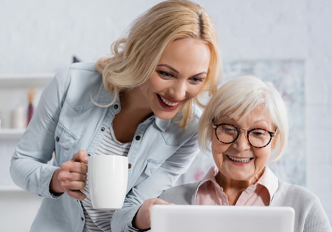 Smiling woman with cup standing near senior parent using digital tablet.